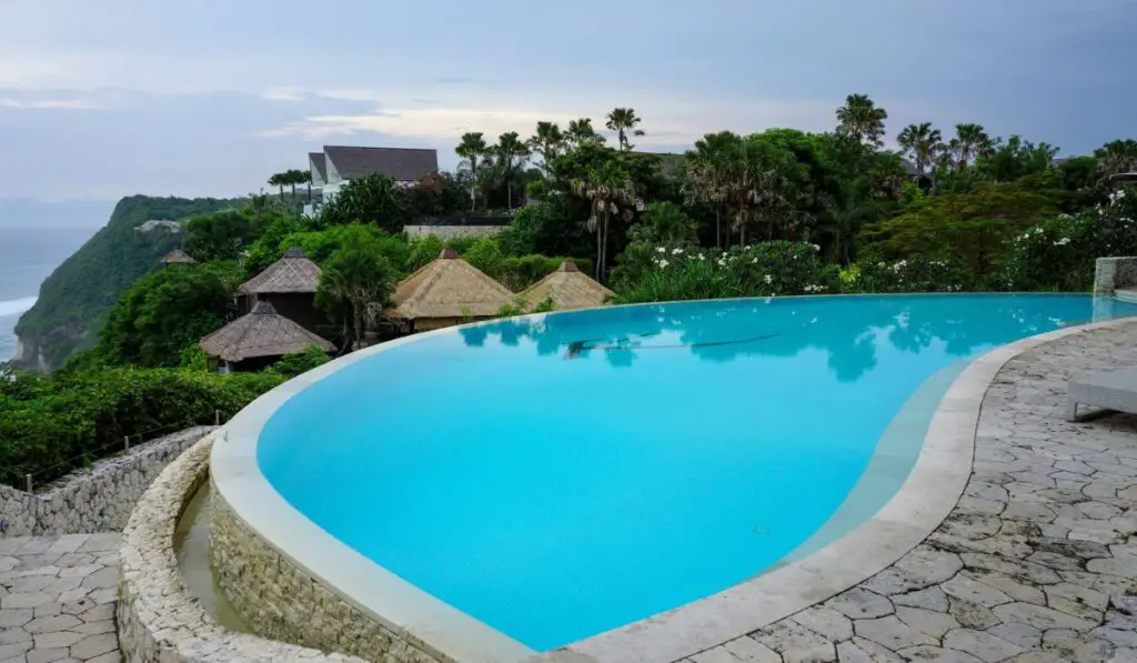 Beautiful view of swimming pool with calm turquoise water in resort with trees and huts in background